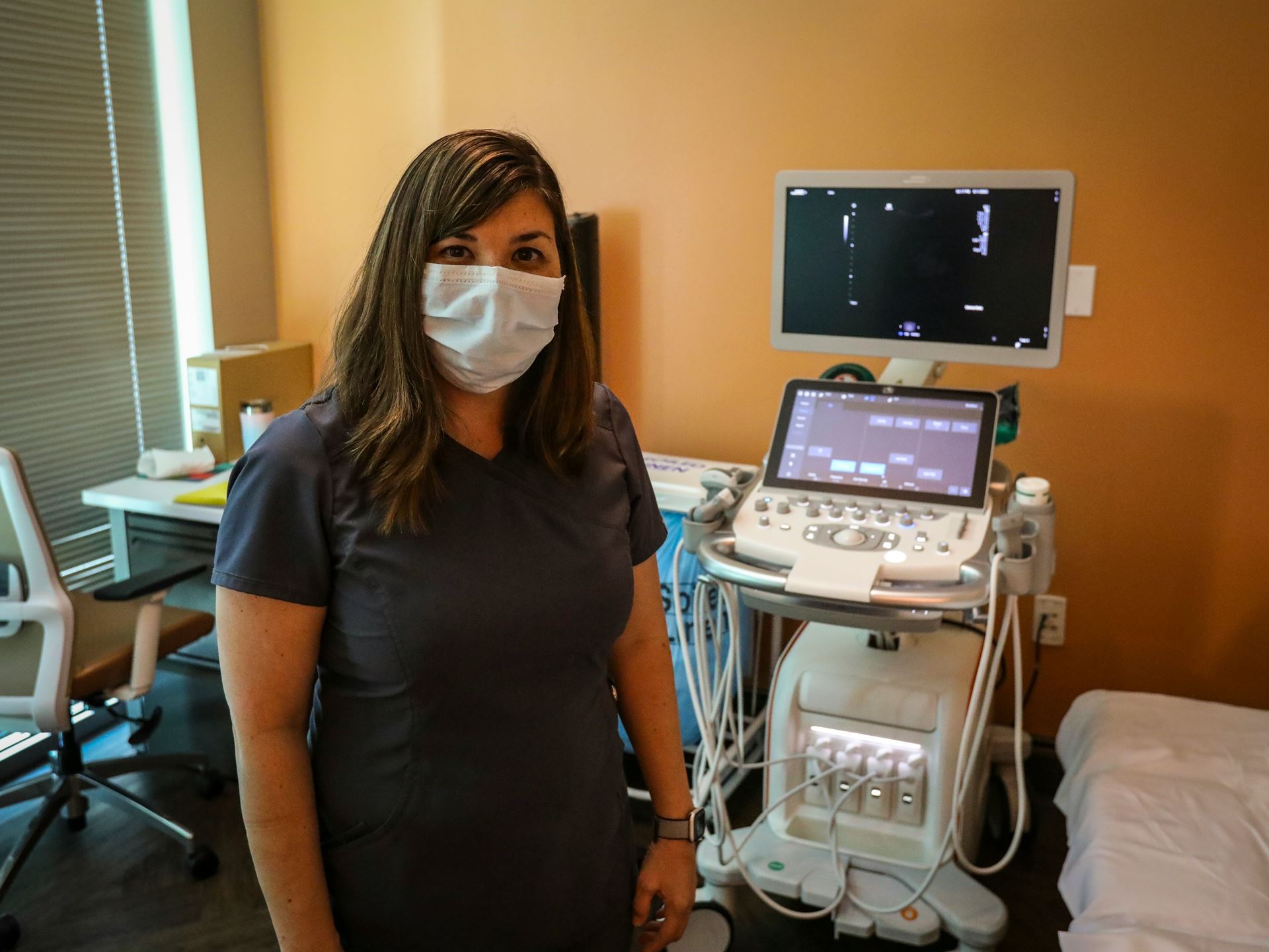 female clinicians in a mask stands in front of an ultrasound scanner